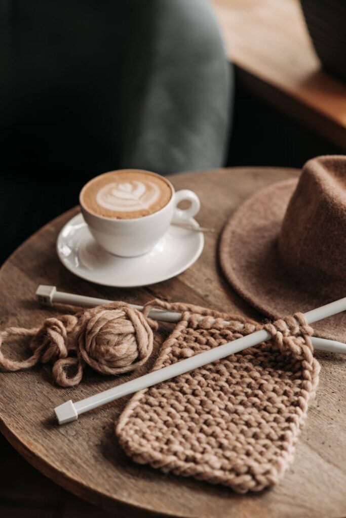 A cup of cappuccino with knitting supplies on a rustic wooden table, conveying comfort and leisure.